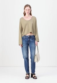 Pulover scurt de culoare verde măsliniu cu mâneci lungi, asortat cu blugi albaștri cu picioare drepte. Modelul ține o geantă de umăr gri deschis și poartă sandale negre.