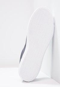 Baskets grises avec une tige synthétique lisse, une semelle en caoutchouc blanche et un motif texturé en zigzag sur la semelle. Vue de côté sur fond blanc.