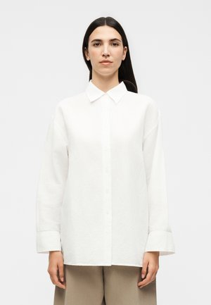 Žena s dlhými tmavými vlasmi v voľnej bielej košeli na gombíky a béžových nohaviciach stojaca proti jednému svetlému pozadiu.