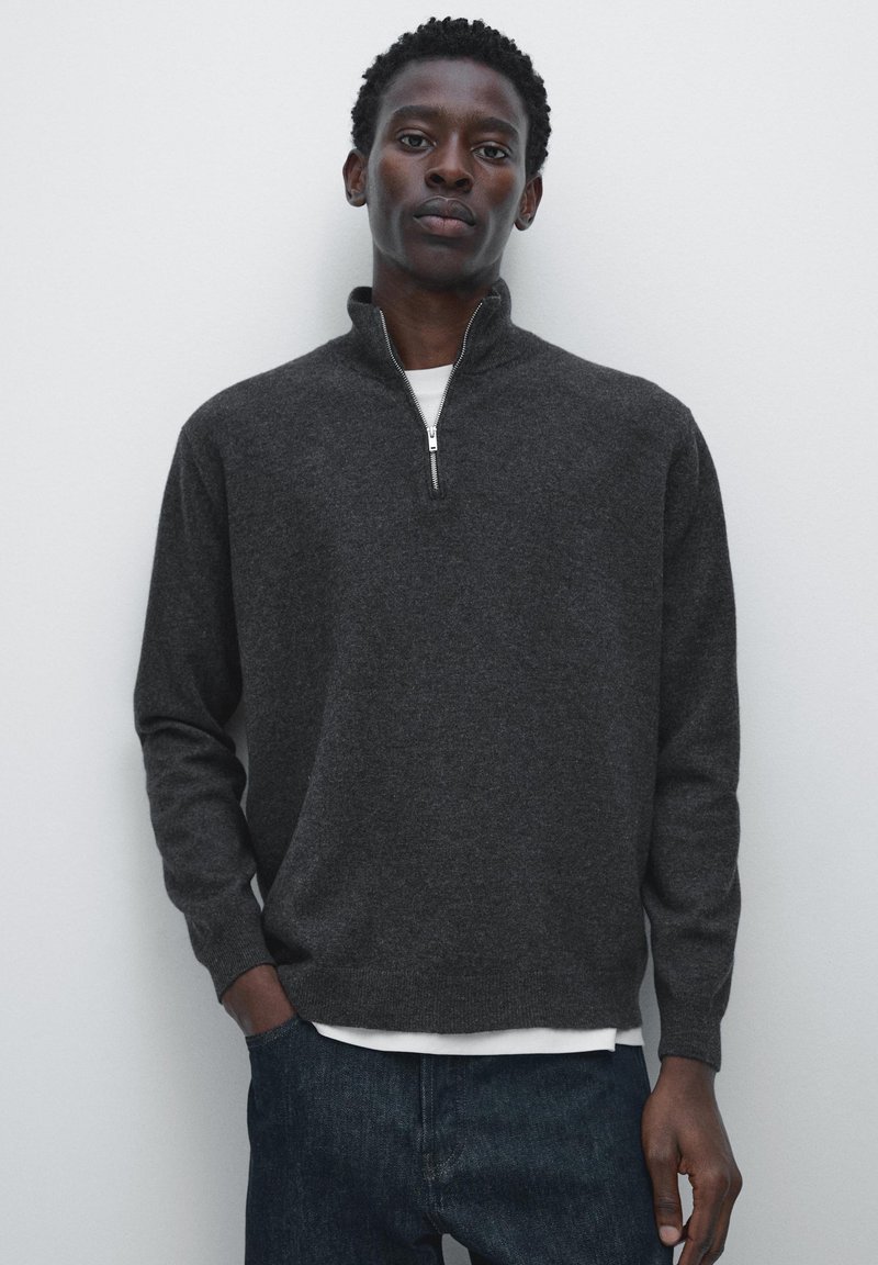Dark grey knit pullover with a quarter zip, long sleeves, and a ribbed hem. Worn over a white shirt with a smooth texture.