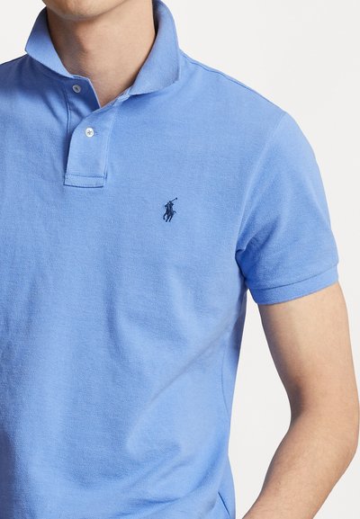 Gaiši zils polo krekls ar apkakli, divām pogām un īsām piedurknēm. Ar mazu, tumšu logotipu kreisajā krūtī. Kokvilnas audums.