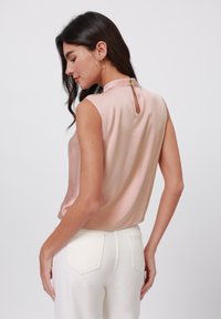 Top sin mangas de color rosa con un escote alto, detalle de abertura en la espalda y textura suave, combinado con pantalones ajustados blancos que muestran los bolsillos traseros.