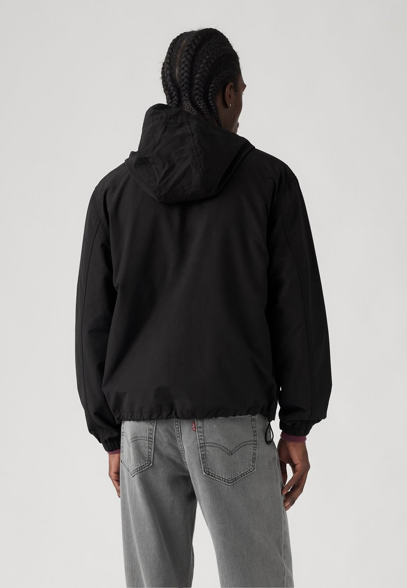 Chaqueta negra con capucha, puños y dobladillo elásticos, que presenta una textura de tejido suave. Combinada con jeans gris claro, que muestran detalles de diseño en la parte posterior.