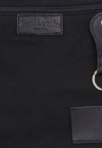 Schwarzer Stoff mit einem aufgenähten Lederetikett mit der Aufschrift "Bulli Leder Studio" und einem Lederband, das einen silbernen Metallring hält.