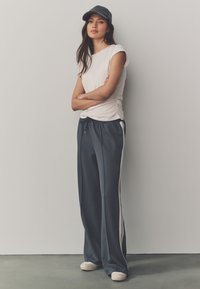 Harmaat leveälahkeiset collegehousut valkoisilla sivuraidoilla, yhdistettynä istuvaan vaaleaan yläosaan. Harmaa lippalakki viimeistelee rennon asun. Karkea kangas.