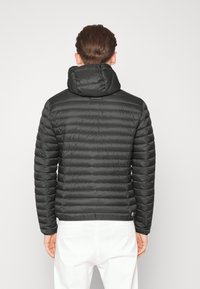 Man i svart vadderad pufferjacka med huva, stående med ryggen mot kameran, i kombination med vita byxor mot en neutral bakgrund.