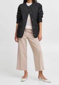 Sort quiltet jakke med trykknapper, med sidelommer, båret over en lys sweater og cremefarvede bukser med vide ben. Hvide spidse flade sko fuldender looket.