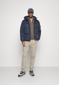 Marineblå pufferjakke med hette, glidelås foran og isolert design, kombinert med en brun genser og beige bukser, fullført med joggesko.