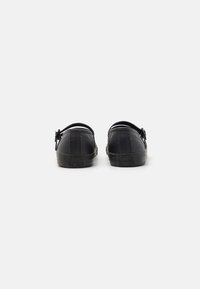 Sapatos pretos de borracha tipo slip-on com bico arredondado, apresentando uma sola texturizada e uma tira traseira ajustável. Branding mínimo no calcanhar.
