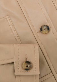 Detailný záber na béžovú látku kabáta, zobrazujúci zapínaný manžetový gombík a gombík na prednej časti s precíznym prešívaním.