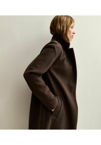 Abrigo de lana marrón con un cuello alto, bolsillos laterales y una textura suave, que presenta una silueta entallada y líneas limpias.