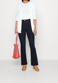 Bílý žebrovaný cardigan přes bílé tričko, tmavé rozšířené džíny, béžové platformové sandály a tkanou tašku červené a bílé barvy.