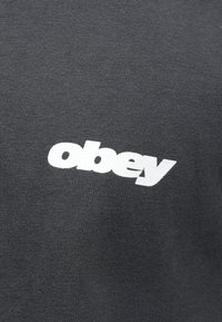 T-shirt de algodão cinza escuro com um destacado logotipo em branco "obey". O tecido é liso com uma textura canelada nas áreas de costura.