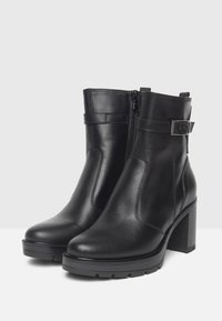 Un par de botas de cuero negro hasta el tobillo con tacones en bloque, cremalleras laterales y correas decorativas con hebillas de metal, sobre un fondo blanco.