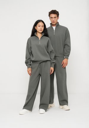 Een vrouw en een man staan naast elkaar, gekleed in bijpassende grijze sweatshirts met ritssluiting en losse broeken met witte sneakers, tegen een effen witte achtergrond.