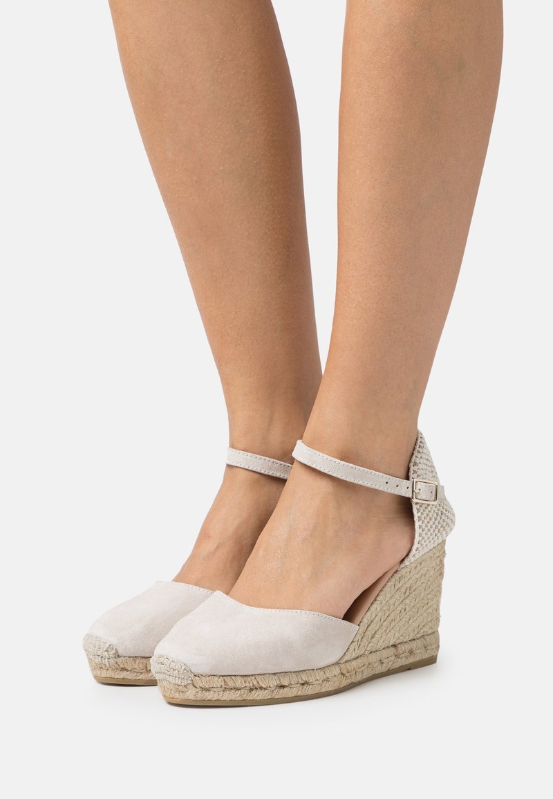 Jambes portant des espadrilles compensées beiges avec des brides à la cheville et des plateformes en jute tissé sur fond blanc.