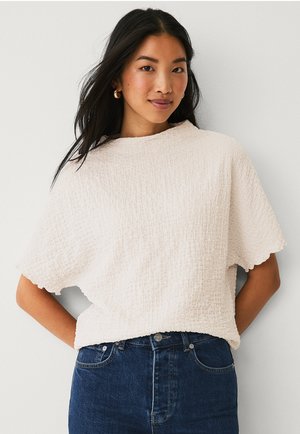 Light beige textured top with a loose fit, short sleeves, and a scalloped hem, paired with dark blue jeans.