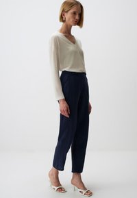 Leichtes Cream-farbenes Langarmshirt mit V-Ausschnitt, kombiniert mit taillierten marineblauen Hosen; Modell trägt weiße offene Sandalen. Schlichtes Design und Passform.