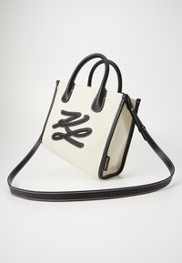 Sac en toile blanc cassé avec des accents en cuir noir, présentant un motif de logo noir, des anses doubles et une bandoulière réglable.