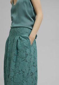 Falda de encaje verde azulado con estampado floral, pliegues y bolsillos laterales. Acompañada de una blusa de satén a juego con un diseño de escote en V.