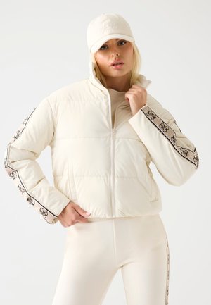 Giacca imbottita bianca con colletto alto e chiusura con zip. Presenta dettagli in contrasto con logo nero sulle maniche. Abbinata a pantaloni attillati coordinati.