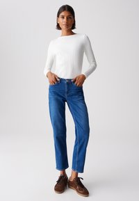 Weißes geripptes Langarmoberteil, hoch taillierte blaue Jeans, braune Wildledersneaker. Schlanker Schnitt mit geraden Beinen, standardmäßige Denim-Textur und Knopfverschluss.