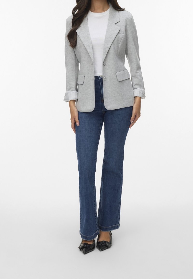 Žena v světle šedém saku přes bílé tričko, modrých džínách a černých lodičkách stojí na bílém pozadí.