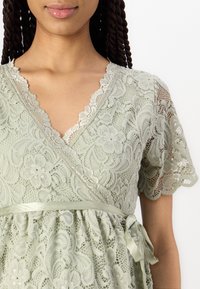 Vaaleanvihreä pitsimekko, jossa on aaltoilevat hihat ja V-pääntie. Sidottu satiinivyö lisää muotoa vyötärölle. Kukkakuvio koko mekossa.