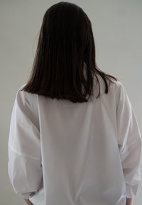 Persoon met schouderlang donker haar die wegkijkt, gekleed in een losse witte langemouwen shirt tegen een effen grijze achtergrond.