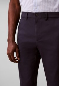 Mann trägt dunkelblaue Anzughose und ein hell gestreiftes Hemd, mit seinem rechten Arm entspannt an der Seite vor einem grauen Hintergrund.