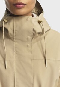 Kvinde iført en beige hættejakke med snoretræk og knaplukning, der viser den nederste del af ansigtet og halsområdet.