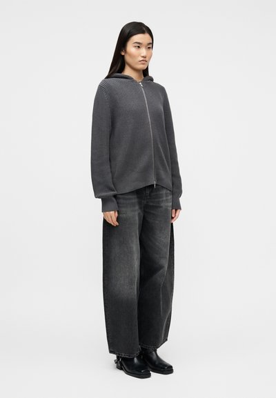 Jeune femme debout portant un sweat à capuche gris en maille avec fermeture éclair, un jean noir délavé à jambes larges, et des bottines en cuir noir.