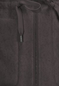 Grau zip-up Kleidungsstück mit Fischgrätenstruktur, ausgestattet mit einem Kordelzug in der Taille und einer seitlichen Reißverschlusstasche. Das Material scheint glatt und strapazierfähig zu sein.