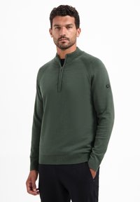 Groene pullovertrui met een hoge kraag, halve ritssluiting, geribde manchetten en onderrand, en een klein logo detail op de linkermouw.