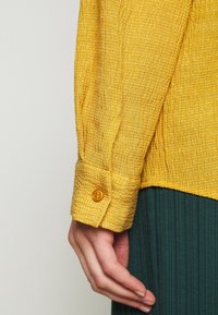 Camicia gialla con texture e polsini abbottonati, caratterizzata da un motivo a griglia. La manica è leggermente arricciata al polso. Il bottone è circolare e giallo.