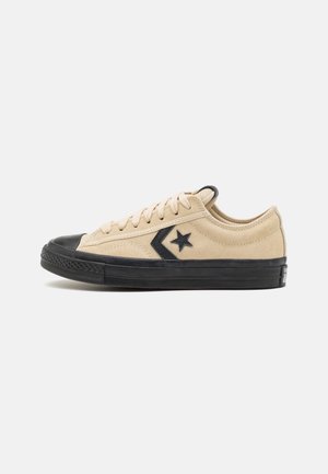 Beżowy zamszowy niskoprofilowy sneaker z czarną gumową podeszwą, czubkiem i logo gwiazdy w kształcie jodełki po boku, przedstawiony na białym tle.