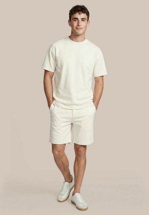 Jonge man staat met handen in de zakken, draagt een crèmekleurig t-shirt, bijpassende korte broek en witte sneakers tegen een effen beige achtergrond.