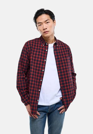 Homme portant une chemise à carreaux rouge et noire boutonnée sur un t-shirt blanc, debout avec les mains près des poches, regardant devant avec une expression neutre.