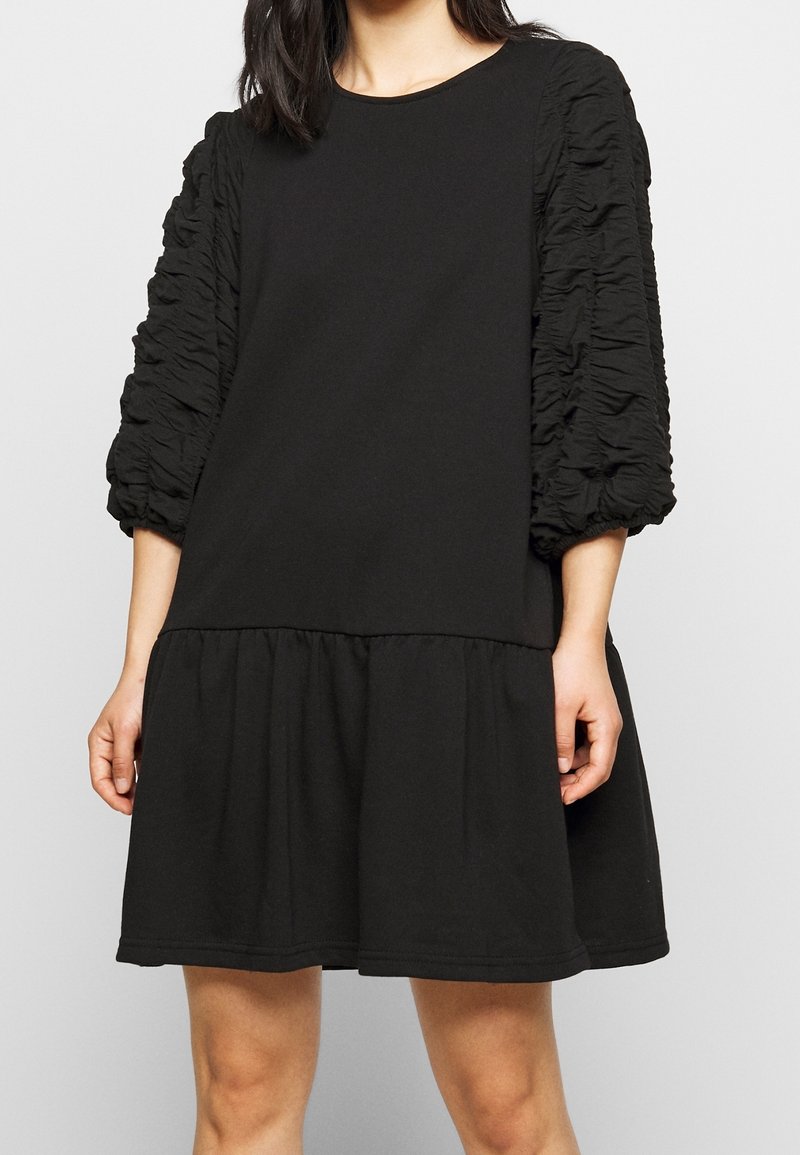 Robe noire avec un corsage ajusté, une jupe à volants en étages et des manches bouffantes ornées d'un design texturé et froncé. Fabriquée en tissu doux.