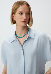 Lichtblauw overhemd met korte mouwen en een kraag, gemaakt van zachte stof. Het model draagt een blauwe en gouden kralenketting die kleurcontrast toevoegt.