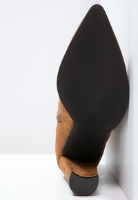 Hnědé semišové kotníkové boty s špičatou špičkou, černou podešví a nízkým podpatkem, postavené v úhlu proti bílé rohu stěny a podlahy.