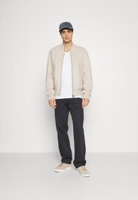Beige bomber jacket, white t-shirt, navy trousers, and beige sneakers. Gray denim cap. Simple, relaxed fit, with a smooth texture.