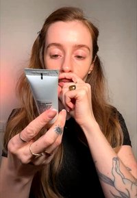 Femme avec des tatouages et des bagues examine le texte sur un tube cosmétique argenté, touchant ses lèvres, sur un fond neutre.