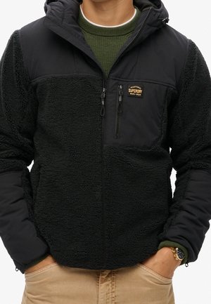 Mann trägt schwarze Fleecejacke mit Reißverschluss und Superdry-Logo, darunter grünen Strickpullover und beige Hose, Hände in den Taschen.