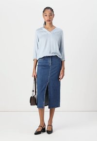 Blusa a quadratini blu chiaro con scollatura a V, abbinata a una gonna di jeans a vita alta con uno spacco frontale. Ballerine nere e una borsa a tracolla marrone.