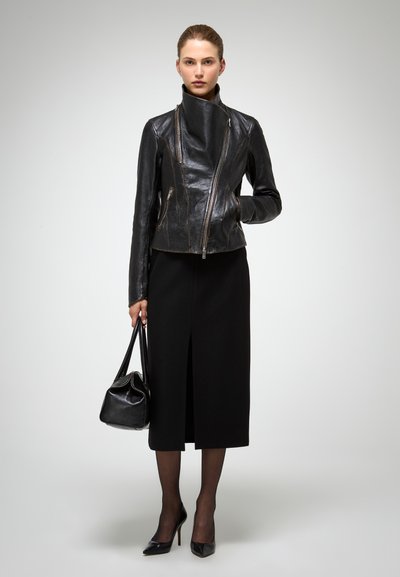 Giacca di pelle nera con zip asimmetrica, colletto alto e due tasche anteriori con zip. Indossata con una lunga gonna nera e tacchi a spillo neri.