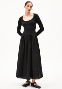 Langes schwarzes Kleid mit einem taillierten, langärmligen Oberteil und gerafftem Rock. Glatte Textur, begleitet von schwarzen Schuhen mit Nietenakzenten.