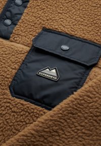 Brown fleece fabric with a smooth navy panel and pocket. Features a logo patch with mountain design. Snap closure for easy access.