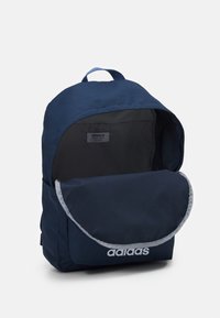 Mörkblå tygryggsäck med rundad topp, grå trim, stor framficka och vit Adidas-logotyp. Insidan är svart med en etikett.