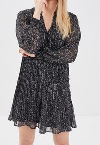 Robe noire plissée avec de longues manches transparentes, ornée d'un motif à pois blancs sur toute la surface et d'une silhouette ample.